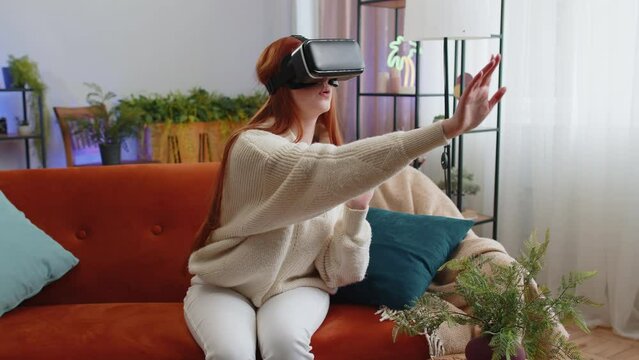 Redhead woman using virtual reality futuristic technology VR app headset helmet to play simulation 3D 360 video game, watching film movie at modern home apartment. Girl in goggles sitting on couch