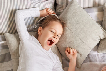 Sleepy little girl yawning in the morning while lying in bed on pillows, keeps mouth open lazy morning on weekend stretching arms with happy facial expression.