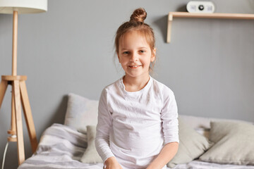 Obraz na płótnie Canvas Positive little girl sitting on bed, smiling kid with bun hairstyle wearing white home clothing looking at camera with optimistic expression, cute emotional child.