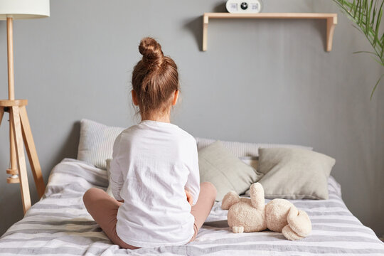 Adorable brown haired girl sitting on bed at bedroom, posing backwards wearing white home clothing, kid with hair bun alone sits on bed.