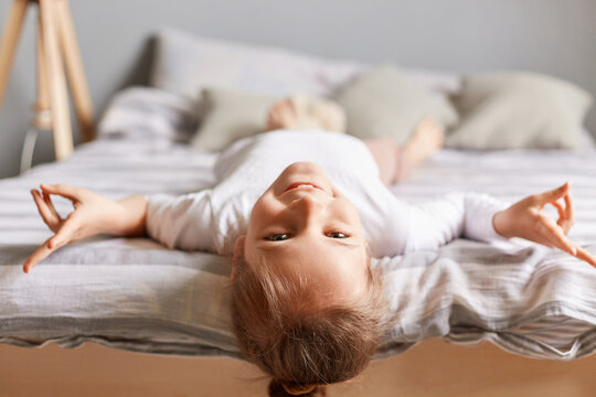 Happy little girl with dark hair lying upside down on bed at home, kid playing having fun showing victory looking smiling to camera.