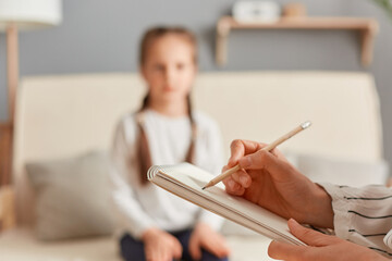 Closeup of doctor psychologist holding paper notebook making notes while talking with small girl at...