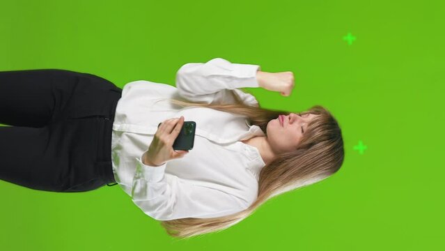 Happy, blonde woman checking smartphone, making winner gesture. Front view of cheerful nice lady with cellphone enjoying, showing hurray sign and dancing, in motion. Concept of happiness, emotions.