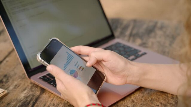 Female freelancer with computer checking statistics charts on smartphone. Woman working remotely on computer checks diagrams on phone. Female analyst studying different graphs. Remote work concept