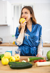 Satisfied woman in dressing gown eating apples in kitchen during breakfast