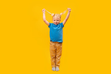 Smiling preschooler wears blue T-shirt on yellow background. Full-length portrait of little blond girl with two ponytails