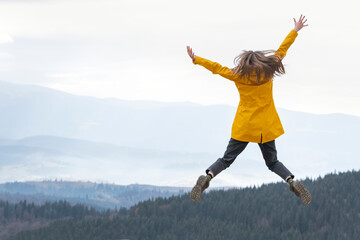 Happy girl in yellow jacket jumps with hands up to the sky on the top of mountains. Travel hiking and tourism.