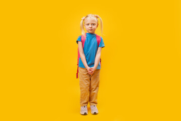 Full-length portrait of elementary school girl with backpack on bright yellow background. Primary school student. Copy space, mockup