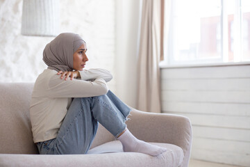 Young woman upset in depression sitting on sofa at home, muslim woman in hijab close up in living room.