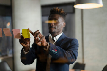 Portrait of successful businessman in office. Young African man witing ideas on colorful stickers on glass wall..