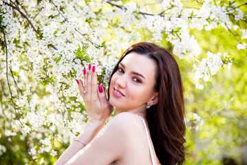 portrait of smiling happy brunette beautiful woman in white T-shirt and pink manicure with white cherry blossoming in the spring sunny day 