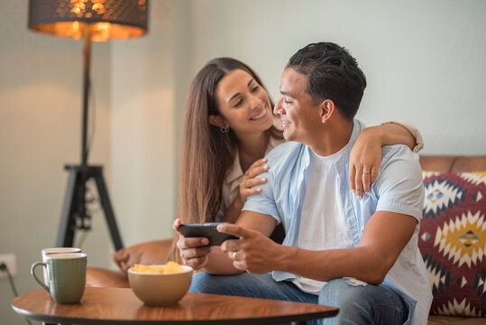 Young couple in real life indoor at home having leisure relax on the sofa and using mobile phone app. Modern young boy and girl living together in apartment. Relationship and dating people lifestyle