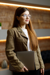 Smiling young woman in glasses and a jacket with a laptop on the background of the office.