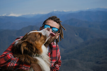 Concept of mountain tourism and travel. Australian Shepherd on hike with owner. Young Caucasian man in sunglasses sitting on hilltop with dog and hugging. Snow capped mountains of the Caucasus range.