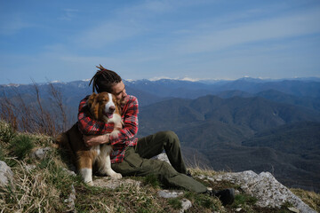 Young Caucasian man with dreadlocks sits on hilltop with dog and hugs. Australian Shepherd on hike...