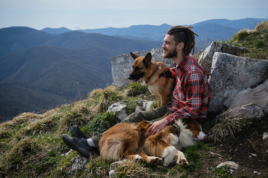 Concept of mountain tourism and travel. Young Caucasian man with dreadlocks sitting on hilltop with two dogs and enjoying views of nature. German and Australian Shepherd travelers resting during hike.