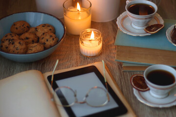 Books, reading glasses, e-reader, plate of chocolate pralines, bowl of cookies, cups of tea and lit candles on the table. Selective focus.