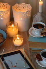 Books, reading glasses, e-reader, plate of chocolate pralines, bowl of cookies, cups of tea and lit candles on the table. Selective focus.