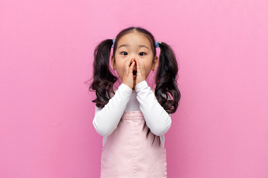 little shocked Asian girl in pink sundress with long hair is surprised and covers her mouth with her hands