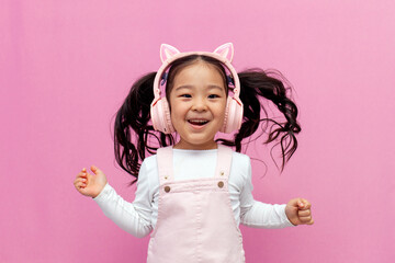 little asian girl in pink children's headphones listens to music and dances on pink isolated background, korean child