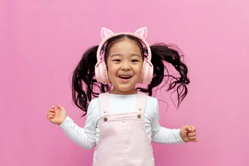 Fotobehang Dansschool little asian girl in pink children's headphones listens to music and dances on pink isolated background, korean child