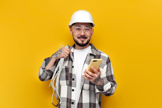 male asian electrician in hard hat and glasses holds internet cable and phone on yellow background, korean worker