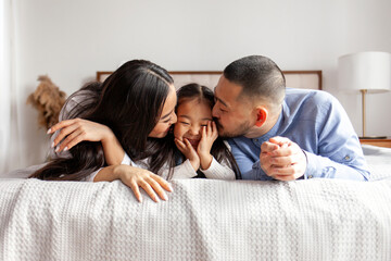 happy young asian family with little daughter lie on bed at home and smile, korean mom and dad kiss...