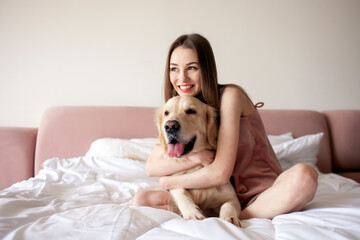 cute girl in pajamas sits on the bed and hugs golden retriever breed dog, woman in the morning at home with pet