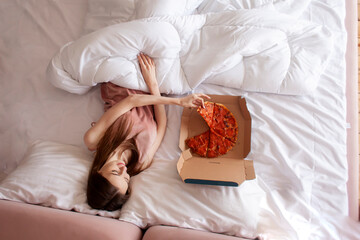 cute woman lies in bed under the covers and eats delicious pizza in the morning, woman in pajamas...