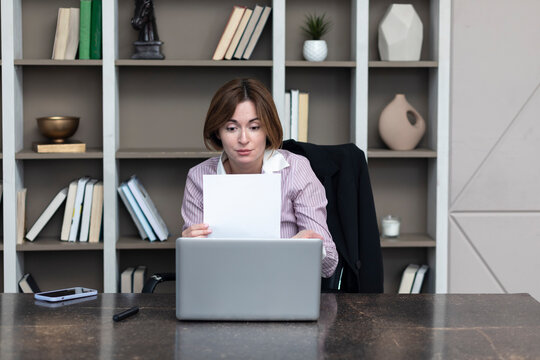 Serious business woman sitting at the desktop, holding documents and talking on video call on laptop in her modern office