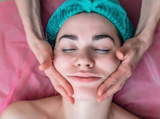 Woman's face in cosmetic cap and hand of cosmetologist. Cosmetic procedures, washing, facial massage