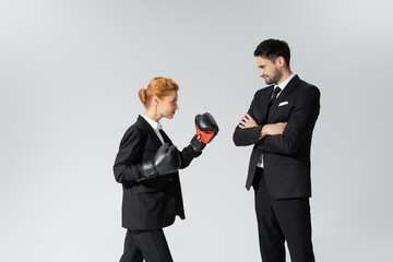 smiling businessman standing with crossed arms and looking at redhead businesswoman in boxing gloves isolated on grey.
