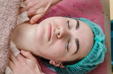Fototapeta na wymiar Woman's face in cosmetic cap and hands of beautician. Cosmetic procedures, washing, facial massage.