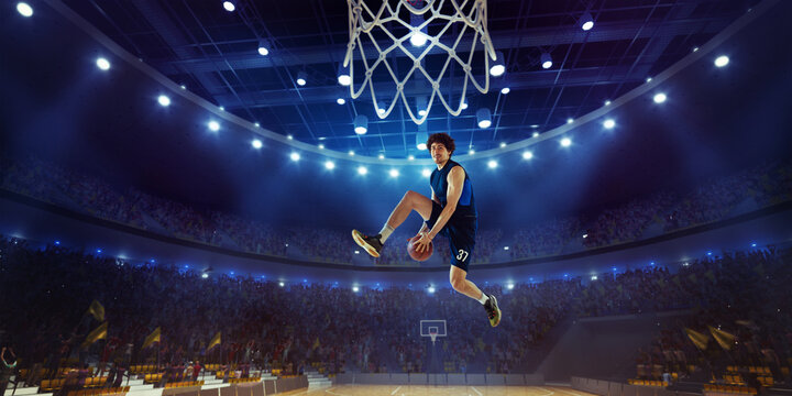 Young man, professional basketball player in motion, jumping with ball during match on 3D stadium with flashlights. Winning game. Concept of professional sport, competition, action and motion