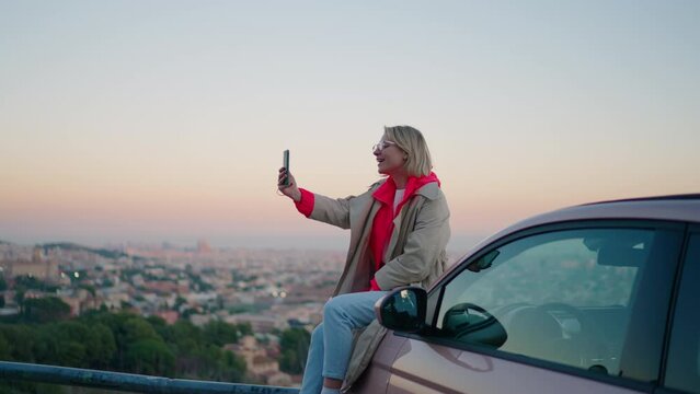 Smiling hipster girl saying Hello while looking at smartphone camera during video call. Attractive millennial female tourist waving and posing to mobile phone while sitting on car during travel 
