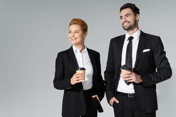 joyful business partners holding coffee to go while standing with hands in pockets and looking away isolated on grey.