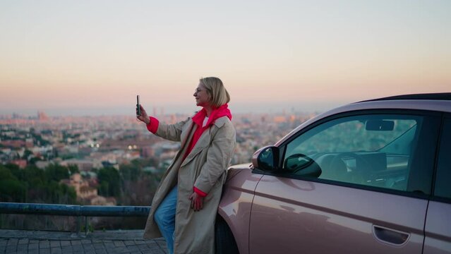 Caucasian smiling hipster girl saying Hello while looking at smartphone camera during video call. Attractive millennial female waving and posing to mobile phone while standing near car during travel