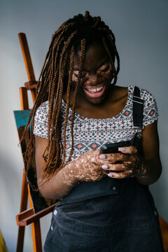 Girl with vitiligo in her painting studio laughing after reading a message on her mobile phone