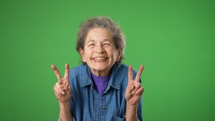 Portrait of happy elderly senior old woman with wrinkled skin and grey hair smiling and giving peace or victory sign gesture with hands isolated on green screen background.