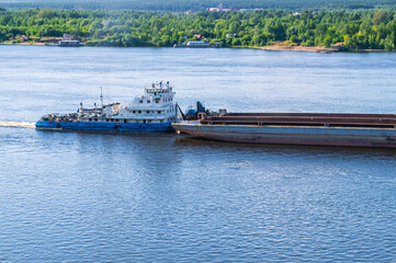 A tugboat pushes a large barge with cargo along a wide river. In summer, the tugboat goes down the...