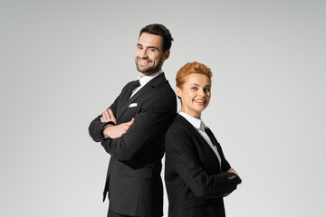 successful corporate managers in black suits standing back to back and smiling at camera isolated on grey.