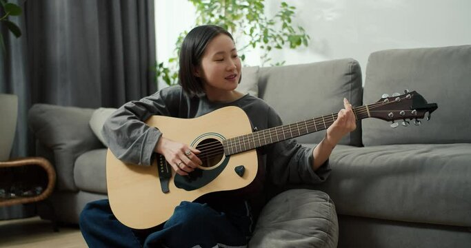 Asian girl playing acoustic guitar music instrument at home, sitting on floor