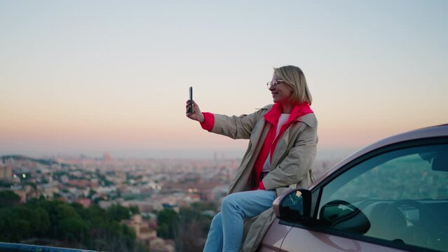 Smiling hipster girl posing at smartphone camera during video call, share experiences and emotions while sits on hood of the car. Female tourist gesturing while communicating via cellular device