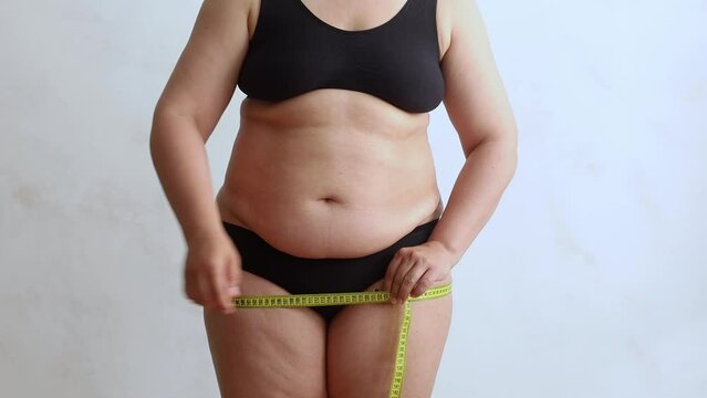 Unrecognizable overweight woman wearing black underwear, measuring buttocks hips with yellow tape, showing result on white background. Weight control, plastic surgery, liposuction, adiposity, diet. 