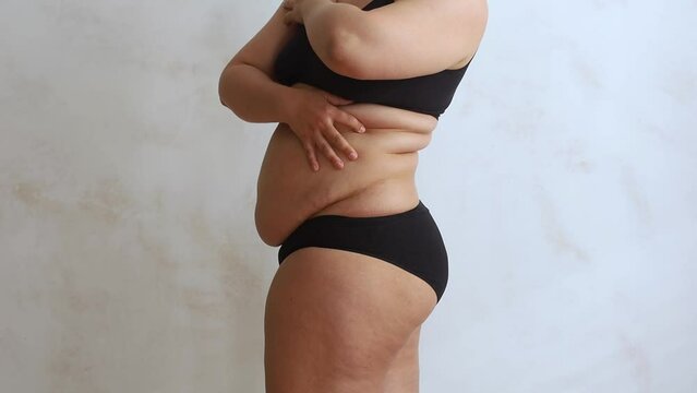 Side view of unrecognizable buxom woman wearing black underwear, lifting excessive fat of stomach with hands, caressing folds on waist on white background. Skin care, weight control, plastic surgery. 
