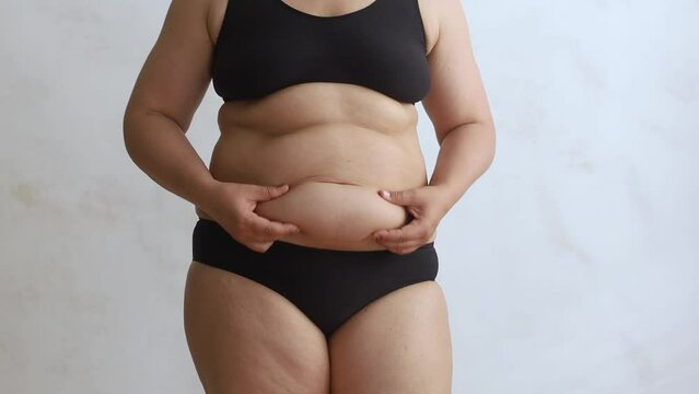 Unrecognizable fat woman wearing black underwear, shaking excessive subcutaneous fat of stomach with hands, doing massage on white background. Weight loss, body care, plastic surgery, liposuction. 