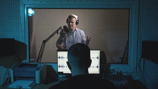a man stands in an isolated booth of a recording studio and speaks into a microphone, while a sound engineer sits at a mixing console. slow motion video .Full HD high quality video recording