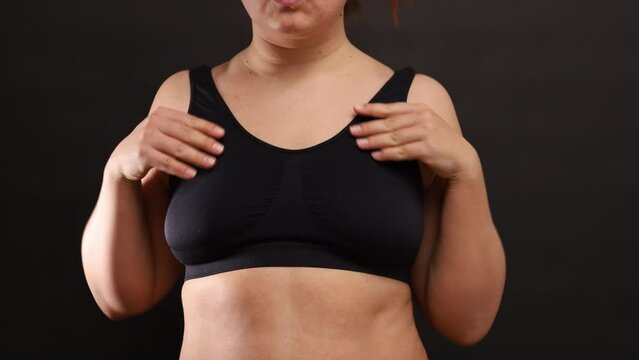 Unrecognizable plump woman adjusting straps, trying on black seamless sports bra, standing on black background, turning side on. Overweight, health, body positive, weight loss, comfortable underwear.
