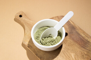 Green powder in a small white ceramic bowl, with a white spoon on a wooden board, on a beige background.  This powder sprinkles the background around.  Super food or marijuana kief concept