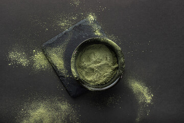 Green powder in a small ceramic bowl, on a stone stand, on a black background.  The background around is sprinkled with this powder.  Super food or marijuana kief concept